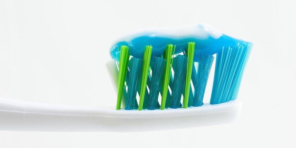 A blue and green toothbrush head with a stripe of blue toothpaste against a white background encouraging you to learn how to brush between teeth